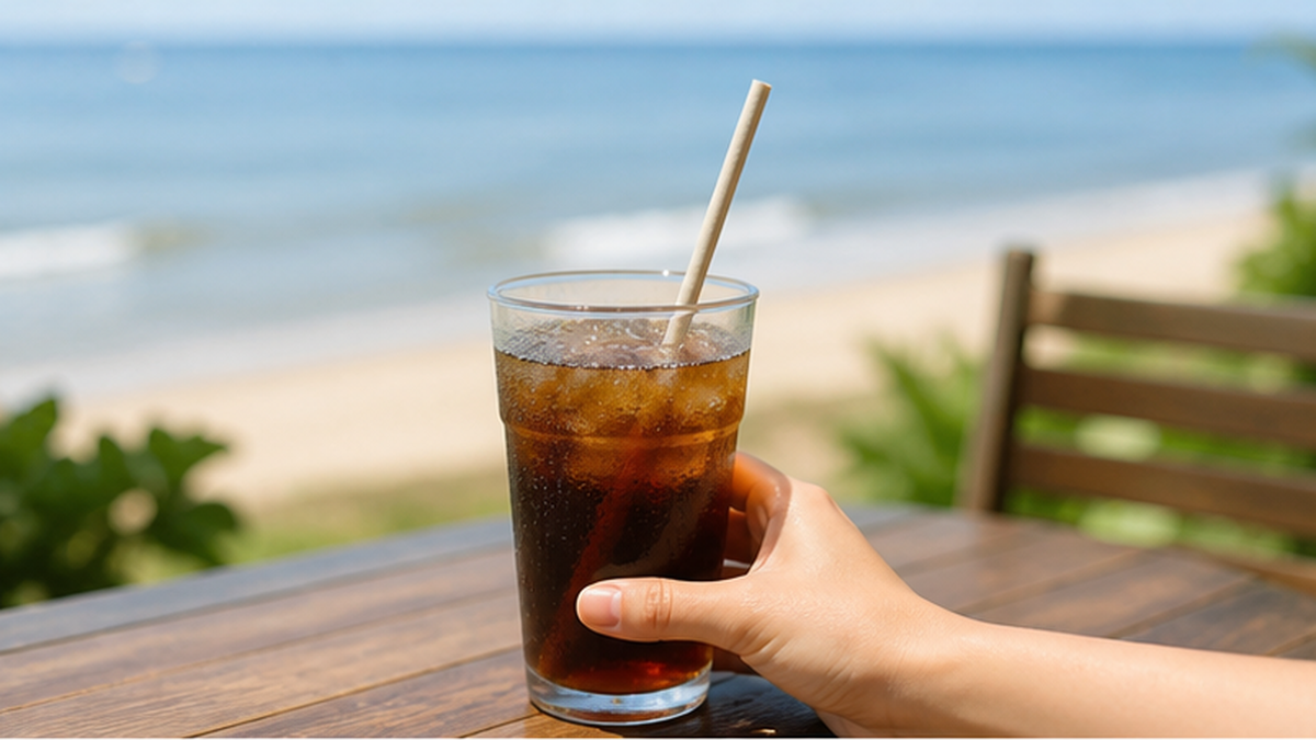 紙ストローが刺さったコーヒーカップを持つ手と、穏やかな海辺の風景